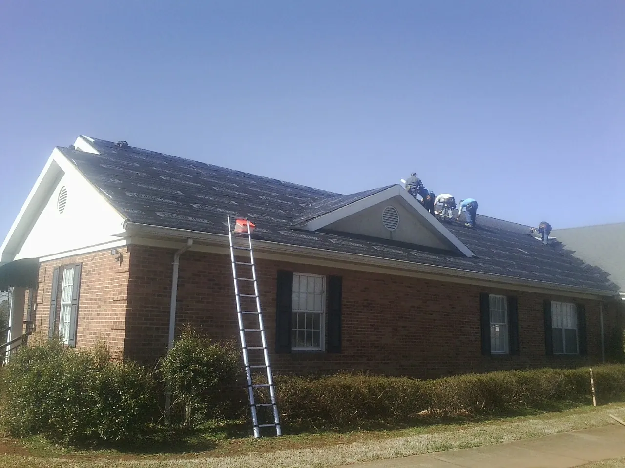 Crew tearing shingles off a brick ranch roof with a ladder set — Metal Roof Repair work by our crew in Duanesburg