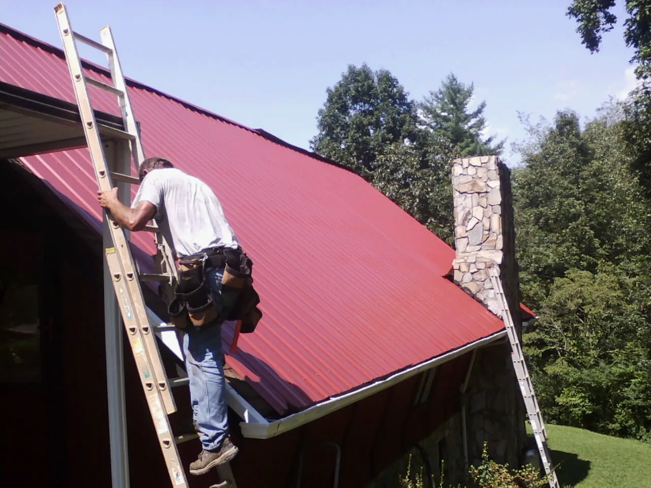 Roofer on a ladder beside a red metal roof and stone chimney — Soffit & Fascia Repair by Roofing Brothers in Duanesburg